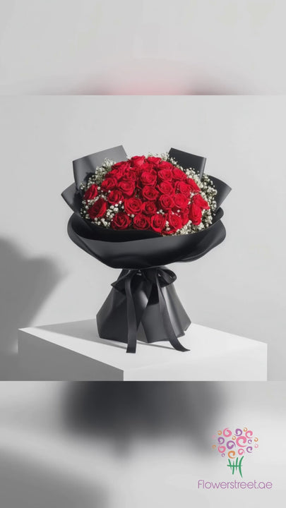 50 red roses with white baby's breath in black wrapping and a black satin ribbon, filmed in warm golden light as rose petals fall past