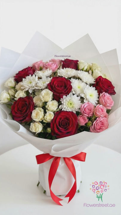 Mixed bouquet of red roses, pink and white spray roses and white chrysanthemums in white wrapping, filmed in warm daylight as petals fall past