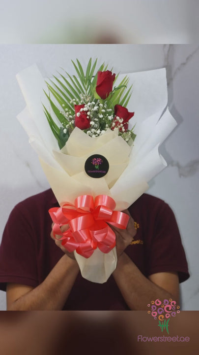 Three red roses with green foliage in white wrapping, filmed in bright daylight as rose petals fall past