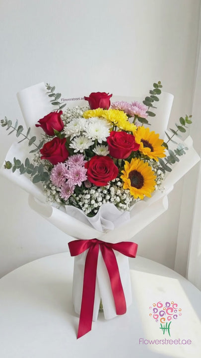 Mixed bouquet of yellow and white chrysanthemums, red roses and a sunflower in white wrapping, filmed in warm daylight as petals fall past